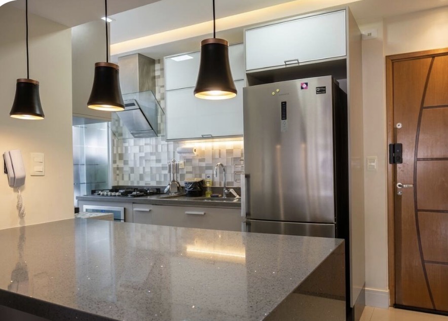 Cozinha com bancada de quartzo cinza, pendentes pretos com detalhe cobre e geladeira inox.